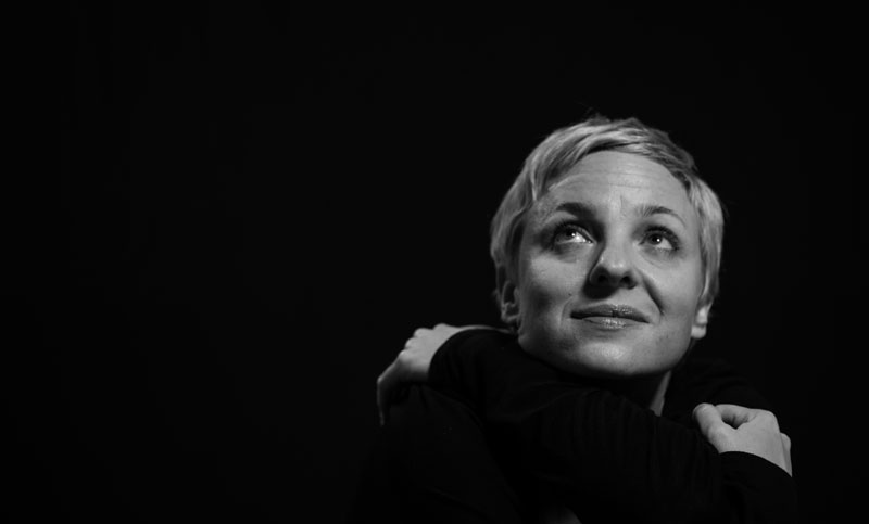 Schwarz-Weiß-Künstlerportrait von Dagmar K. mit sehr kurzem blonden Haar, die verträumt lächelnd sich selbst umarmt und aus dem Schwarz zur Decke schaut.